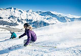 Two skiers carving turns in the powder snow of the Lenzerheide ski area with an impressive mountain panorama and brilliant blue sky.